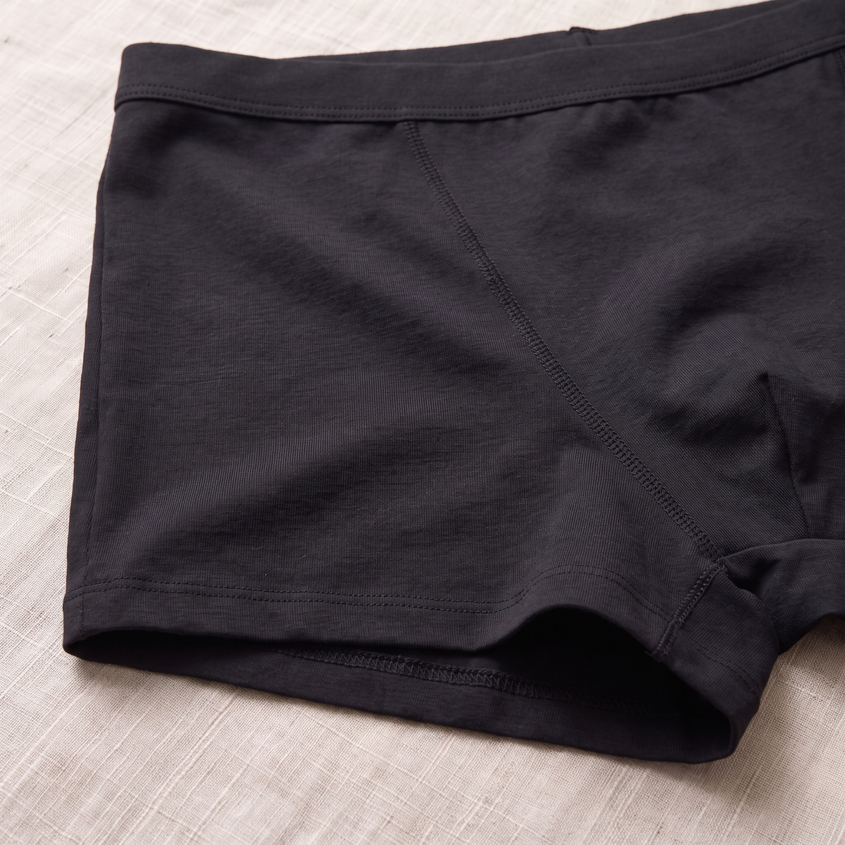 Close-up of a pair of black men's boxer briefs laid flat on a light beige textured fabric background.