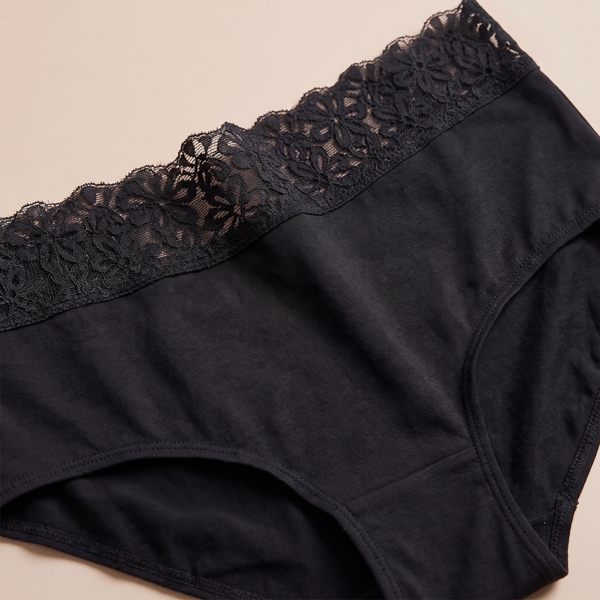 Close-up of black underwear with a wide floral lace waistband, photographed on a plain light beige background.