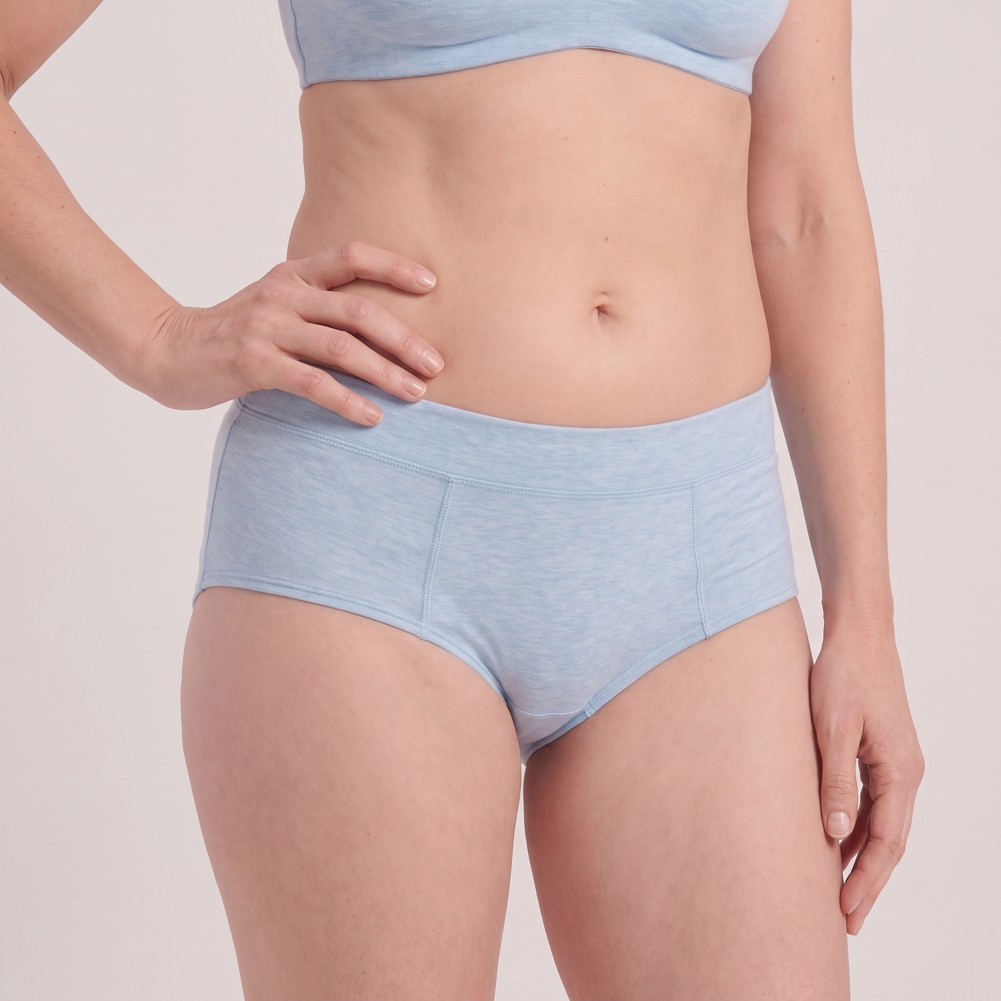 A close-up of a model wearing light blue high-waisted briefs and a matching light blue sports bra against a solid pale beige background.
