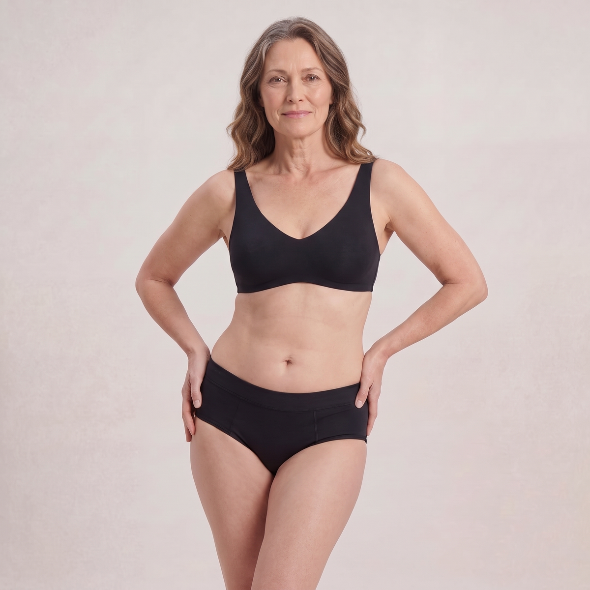 A woman modeling a matching black bra and underwear set against a plain, light beige studio background.