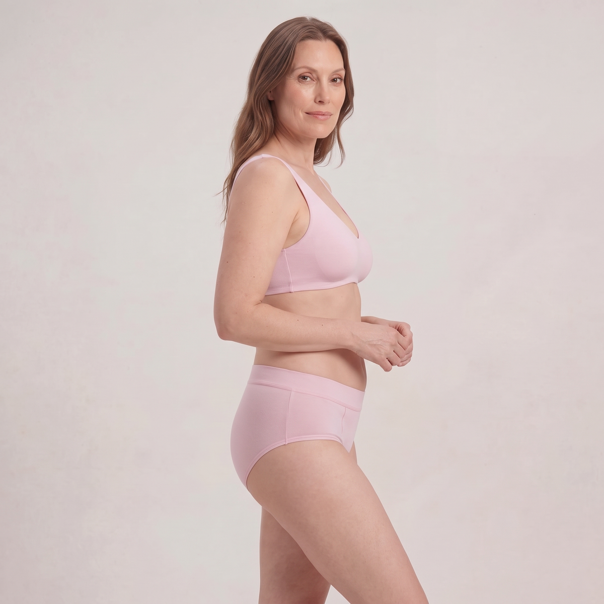 A woman modeling a light pink bra and matching high-waisted briefs against a plain, light beige studio background.