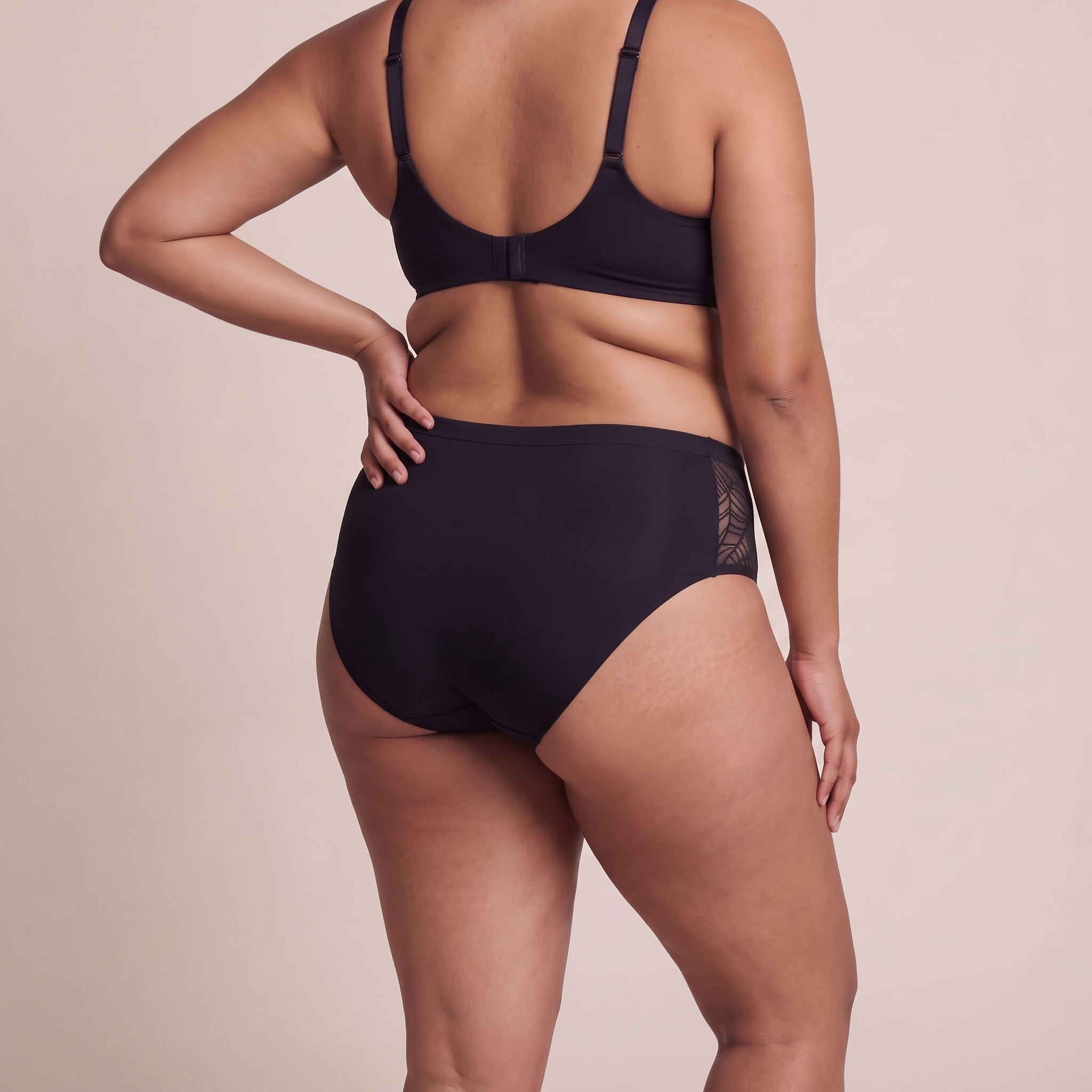 A model shown from the back wearing a black bra and matching high-waisted briefs with lace side panels, set against a solid beige studio background.
