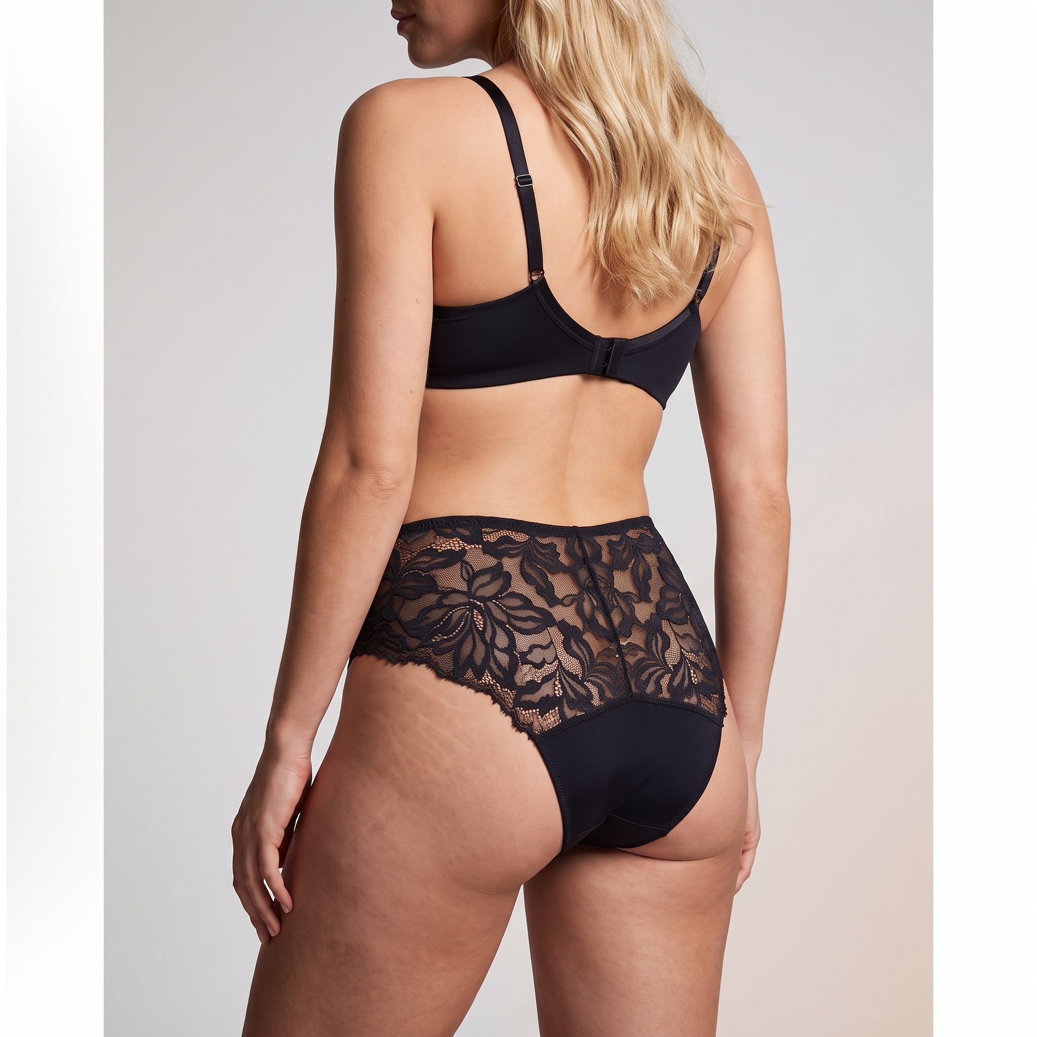 A woman shown from the back wearing a black bra and matching high-waisted black lace panties against a plain light gray studio background.